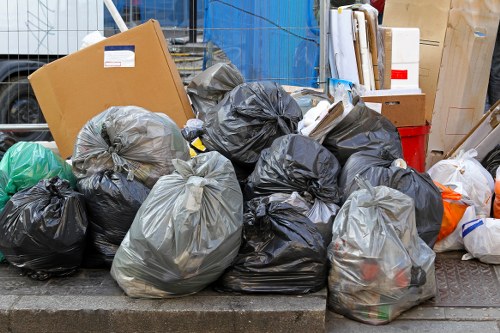A collection of black and grey plastic rubbish bags, tightly tied at the top, are piled on a paved surface with some stacked on each other and others leaning against the curb, indicating an outdoor waste collection. Among the bags, visible items include a cardboard box, plastic bottles, and miscellaneous household waste, suggesting clutter from garden or home clearance. The bags are situated in a residential or commercial area, with background elements such as a blue construction fence, a red plastic bin, and some wooden fencing or paneling partially visible. The overall scene depicts a typical rubbish disposal setup, which Garden Clearance Queens Park likely manages, with the area prepared for collection or removal of garden or household waste in the Queens Park neighbourhood. The lighting is natural, with shadows indicating daylight, and the environment appears organized for rubbish disposal and collection purposes.