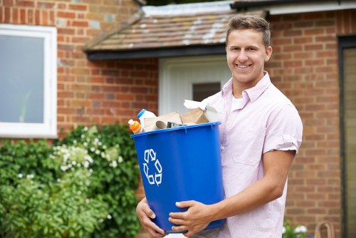 A young man with light brown hair smiling and wearing a light pink short-sleeved shirt, standing outdoors in front of a red brick house with a white-framed window. He is holding a large blue recycling bin filled with various types of rubbish, including brown paper bags, plastic bottles, and other waste materials. The bin features a prominent recycling symbol in white. In the background, there are green bushes and flowering plants, with a paved driveway or pathway visible near the house. The scene appears to be during daytime with natural lighting, illustrating a typical rubbish collection or disposal activity related to garden or household waste management in Queens Park or nearby areas, consistent with the services offered by Garden Clearance Queens Park.