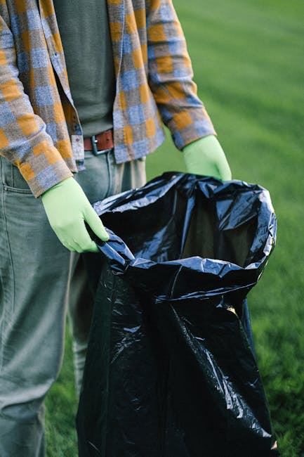 A person wearing a plaid shirt, beige trousers, and bright green gloves is holding open a large black rubbish bag outdoors, ready for waste collection. The bag appears to be made of plastic with a glo