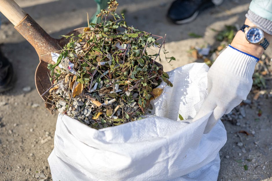 A person wearing a plaid shirt, beige trousers, and bright green gloves is holding open a large black rubbish bag outdoors, ready for waste collection. The bag appears to be made of plastic with a glo